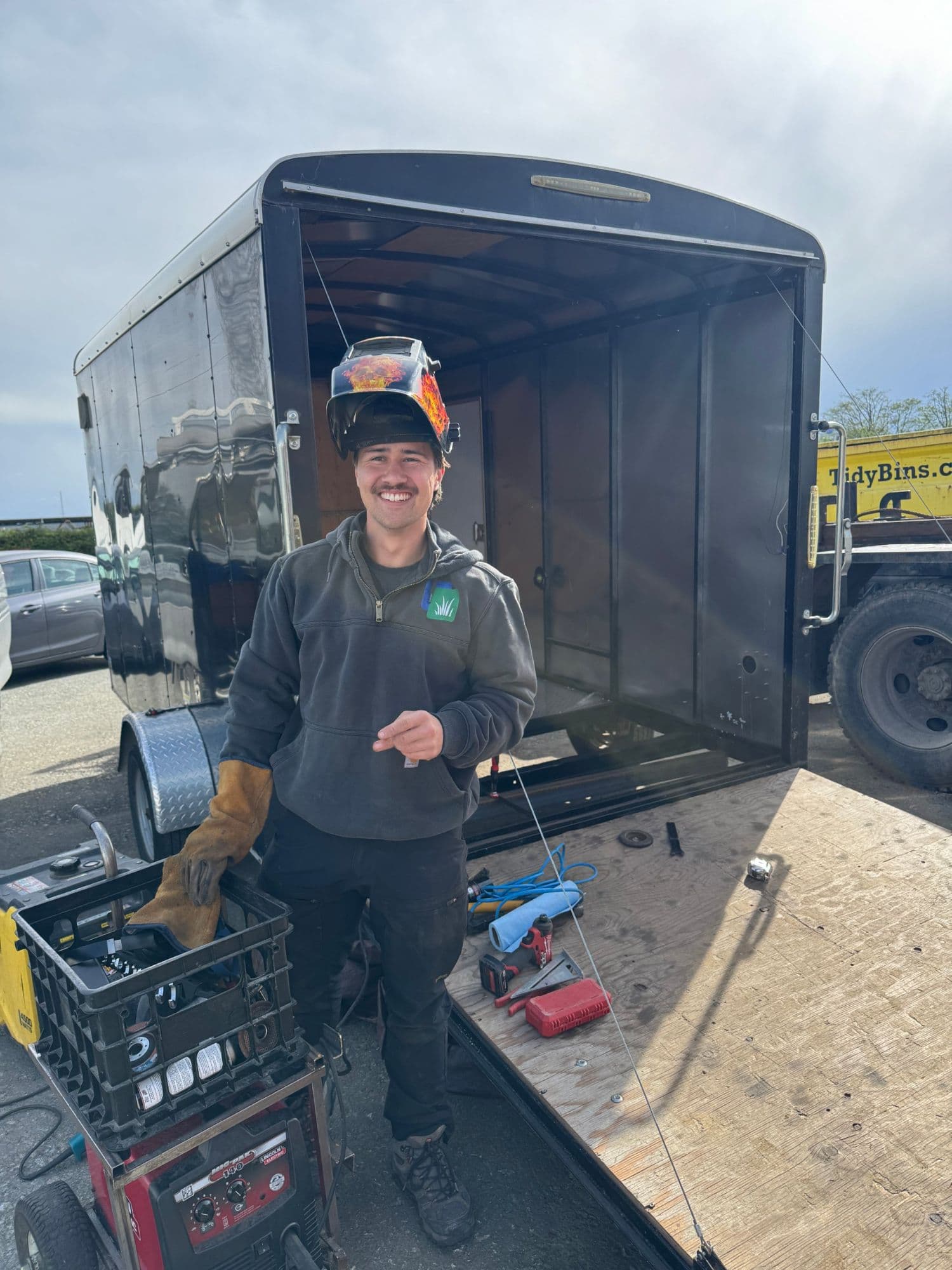 Julian, Kyst’s creator, smiling with a welding helmet pushed up on his head, standing in front of an open black trailer with tools around him